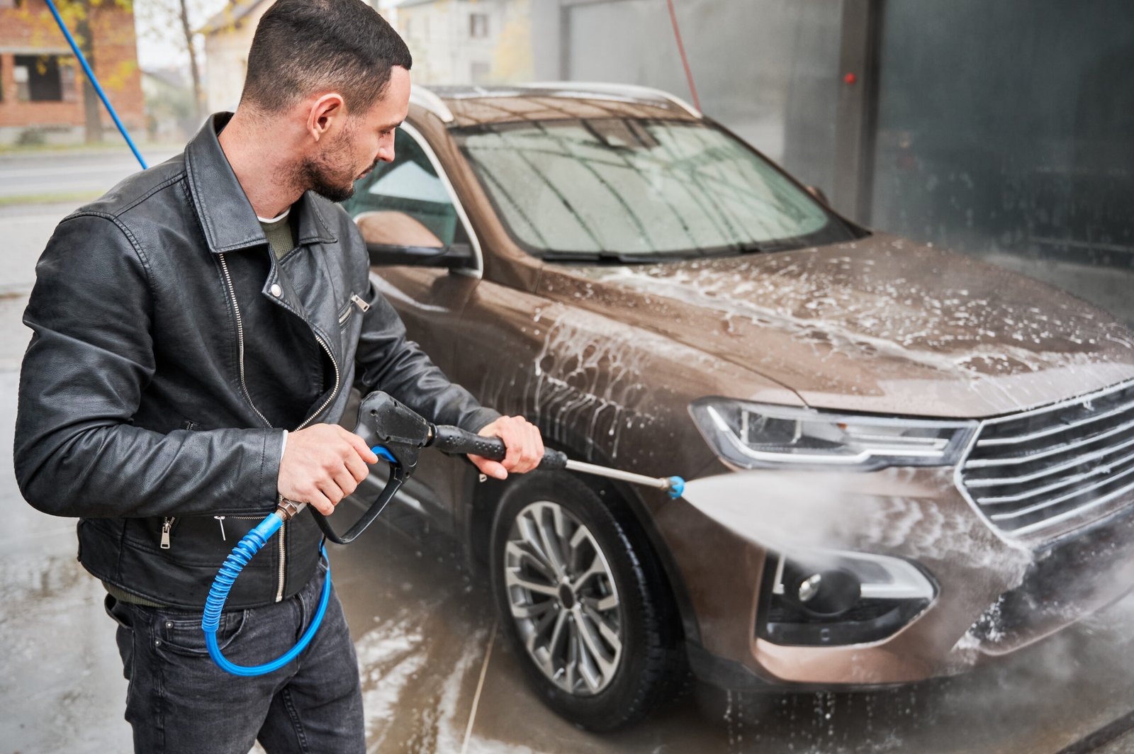 Process of detailing car cleaning. Guy in casual clothes spraying water under high pressure on his automobile, using water gun. Non-contact cleaning of auto body at car wash.